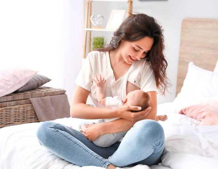 Uma mãe sorridente amamentando o seu bebê com leite materno, sentada em sua cama.