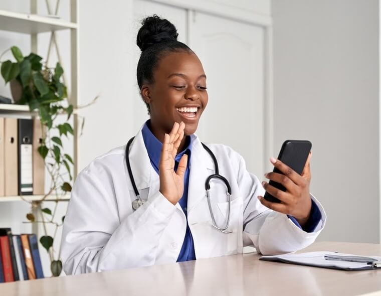 Médica negra fazendo uma teleconsulta (consulta online) pelo celular e acenando para o paciente.